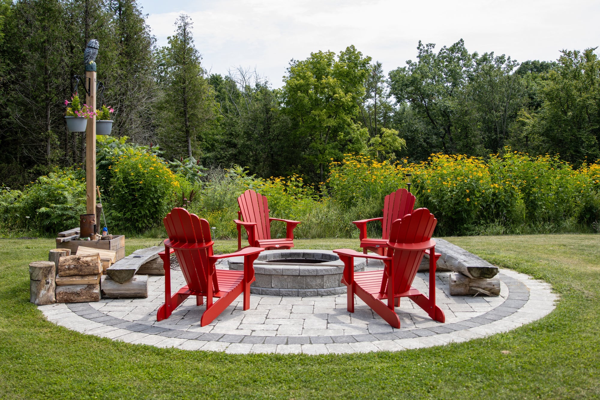 Circular paver fire pit lounge with Adirondack chairs, lawn, and a flowering garden backdrop by Elzo Landscape